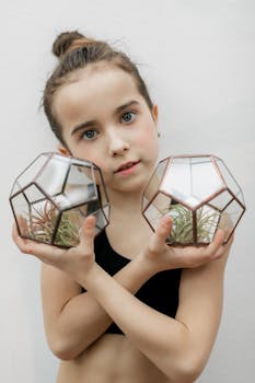 A young girl holds two geometric glass terrariums with plants inside on a white background.