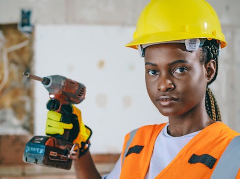 Portrait of a handywoman wearing a hardhat and reflective vest, holding a drill in a construction setting.